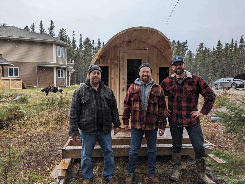 Three men standing in front of a wooden cabin in a rural setting.